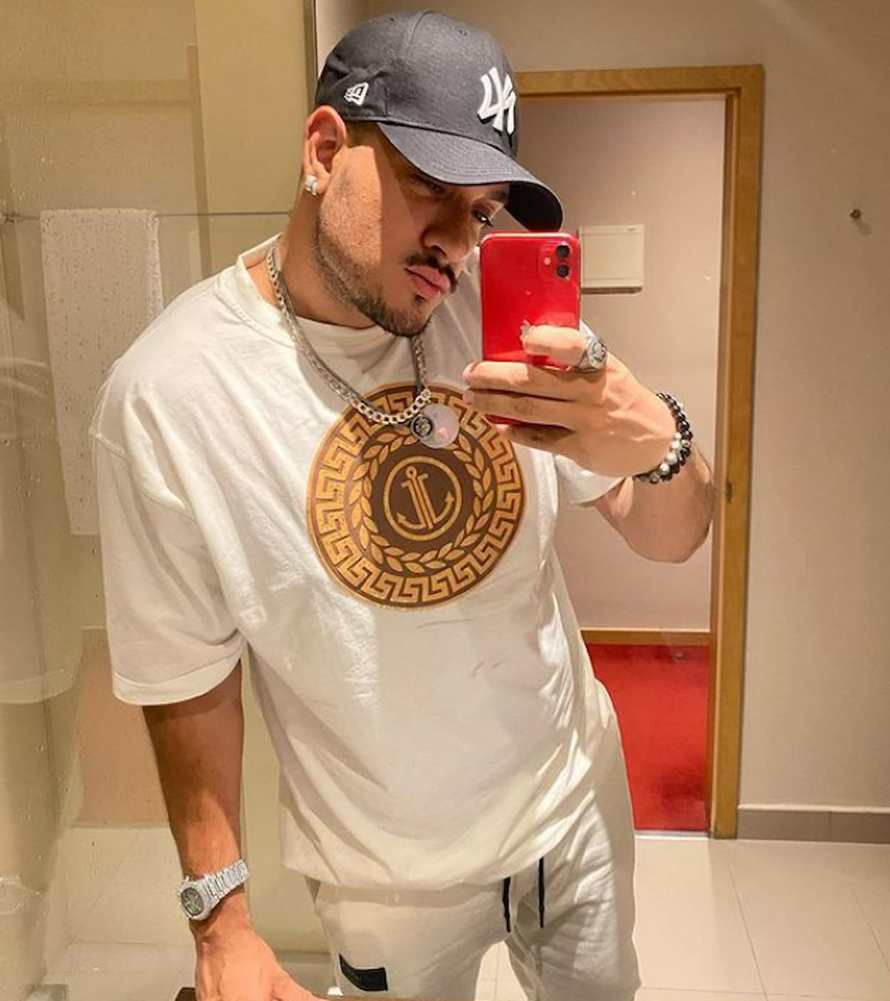 MC Bin Laden tirando foto no espelho com celular, usa boné, camiseta com círculo, relógio e colares
