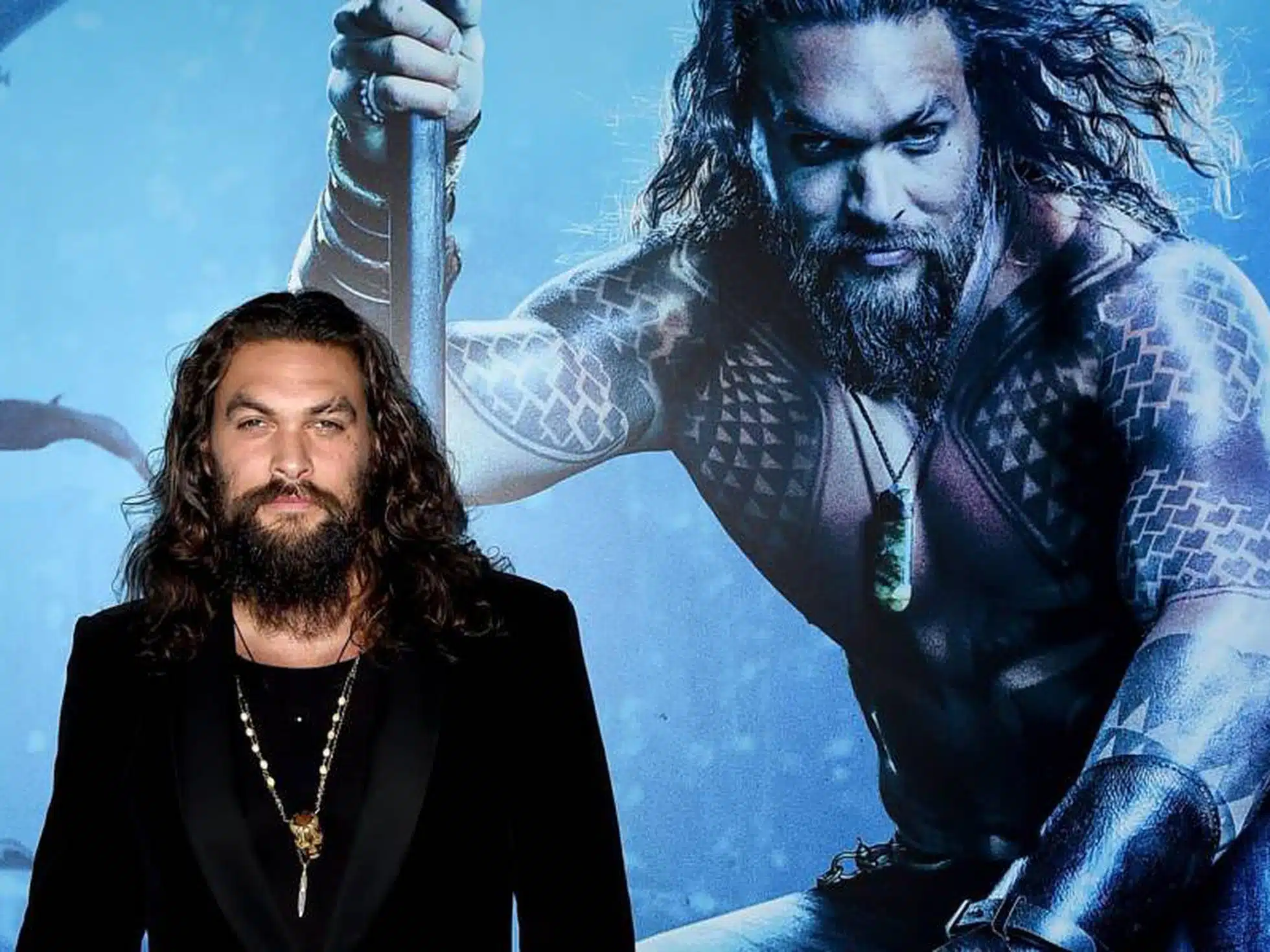 Imagem mostrando o ator Jason Momoa ao lado do cartaz de seu filme Aquaman